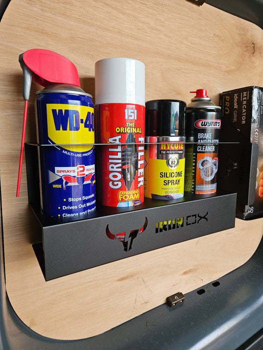 A black shelf, designed as an Aerosol Can Holder, holds five cans: WD-40, Gorilla Filler, CRC Silicone Spray, Wynn's Brake and Parts Cleaner, and another cleaner. Its wall-mounted design is ideal for van & workshop storage against a wooden surface.