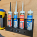 A wall-mounted metal shelf, boasting high-quality construction, holds four tubes of filler and silicone sealant in various colors, alongside a container of blue cleaner. Its space-saving design ensures an organized and efficient workspace. This is the Silicone Mastic Holder.