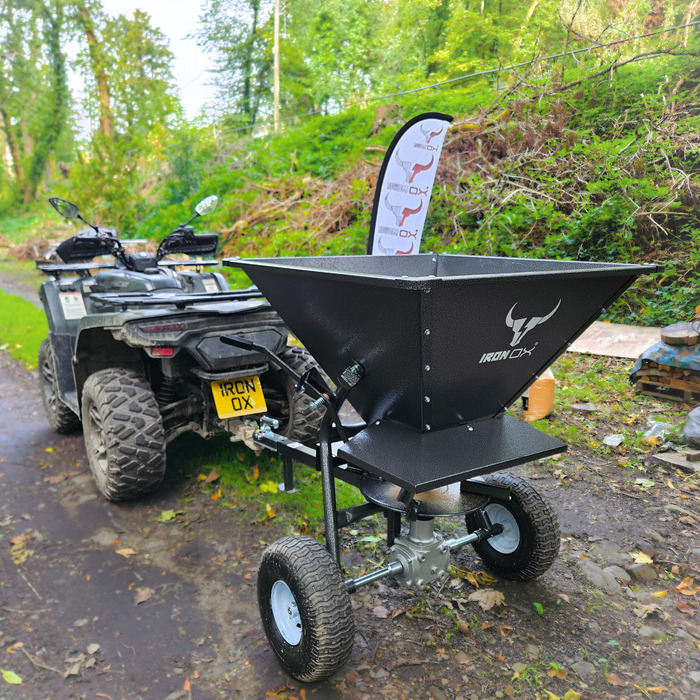 An all-terrain vehicle with a Towable Spreader attached stands on a forest path, promising effortless precision.