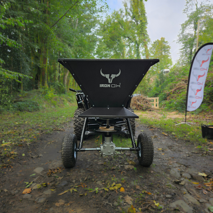 A black Towable Spreader with large back wheels and a triangular attachment is positioned on a forest path, surrounded by greenery and a company banner to the right.