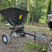 The Towable Spreader, an ATV-towed device with a durable black hopper for distributing materials, is hitched to a vehicle in a wooded area. This device ensures effortless precision in all your landscaping tasks.