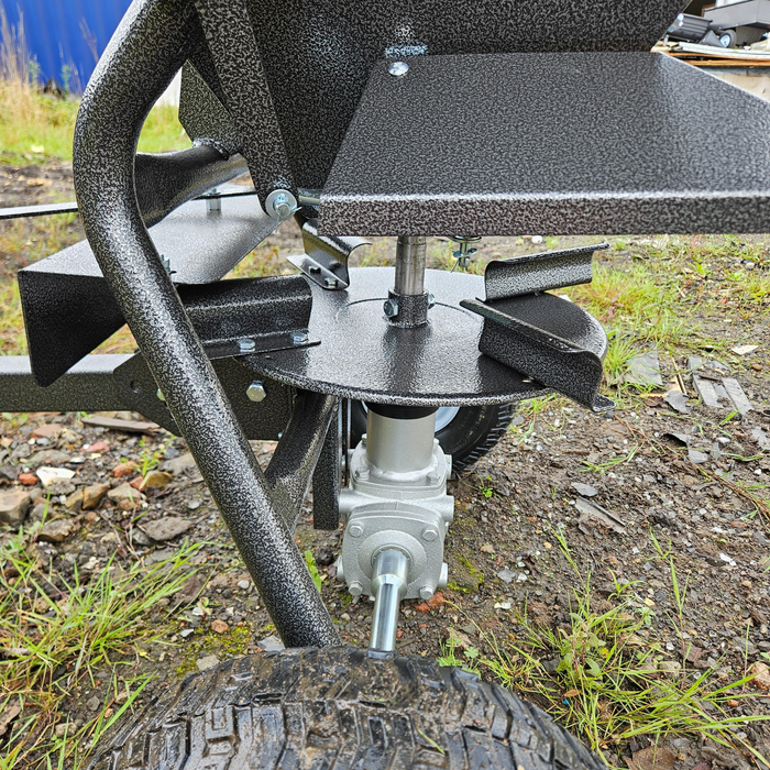 Close-up of a metal agricultural machine mechanism with large black tires, displayed on a gravel and grass-covered ground—the Towable Spreader exemplifies effortless precision.