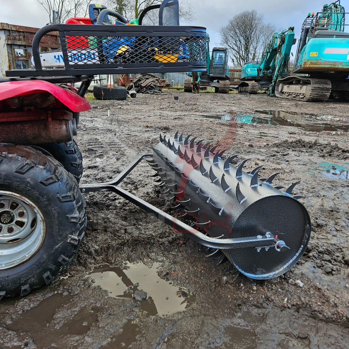 A robust Towable Spiked Roller Aerator 60" Wide is connected to an ATV on muddy terrain, with construction machinery in the background, guaranteeing effective aeration.